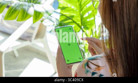 Asiatische Frau mit Brille mit Finger für Touch-on-Phone Mockup mit leerem grünen Bildschirm mit Kopierraum in der Hand, schauen Sie sich das Smartphone zu Hause an. Stockfoto