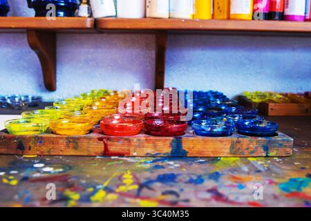 Glasschüsseln mit leuchtenden gelben, roten und blauen Pigmenten sind ordentlich in Reihen auf einem kunstvoll bespritzten Holztisch angeordnet, auf dem weitere Kunstgegenstände stehen Stockfoto