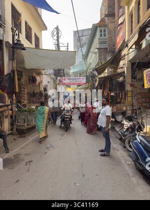 Straßenszene in einem Viertel mit Menschen, Motorrädern und Geschäften bei sonnigem Wetter, Varanasi, Uttar Pradesh, Indien Stockfoto