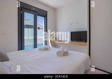 Schlafzimmer mit Sonnenlicht, Doppelbett mit frischer Bettwäsche, Wandschreibtisch mit Fernseher und Blick auf den privaten Pool durch große Glastüren. Stockfoto