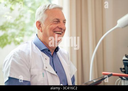 Kopfaufnahme eines fröhlichen und erfahrenen männlichen Arztes, der während einer Konsultation in seinem modernen Büro lacht und ein digitales Tablet hält. Stockfoto