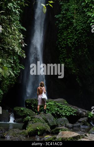 Frau, die auf Felsen unter dem schmalen Wasserfall im tropischen Wald von Bali steht Stockfoto