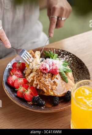 Nahaufnahme einer Person, die einen Stapel Pfannkuchen mit frischen Beeren und Sahne mit einer Gabel und einem Messer schneidet. Gemütliche Frühstücks- oder Brunch-Szene. Stockfoto