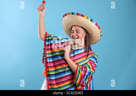 Glückliche Frau in Sombrero-Hut und farbenfrohem Poncho, der Maracas auf hellblauem Hintergrund spielt Stockfoto