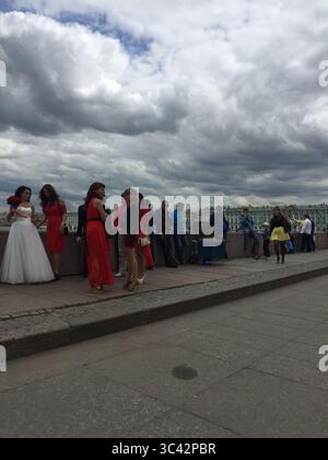 Eine Gruppe von Menschen, darunter eine Braut in weiß und andere in roten Kleidern, versammeln sich auf einer Uferpromenade in Sankt Petersburg unter einem bewölkten Himmel Stockfoto
