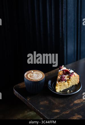 Ein Stück Kuchen, serviert mit einer Tasse Cappuccino mit Latte Art, auf einem rustikalen Holztisch. Gemütliche und einladende Café-Szene, perfekt für einen Rela Stockfoto
