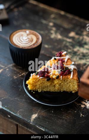 Ein Stück Kuchen, serviert mit einer Tasse Cappuccino mit Latte Art, auf einem rustikalen Holztisch. Gemütliche und einladende Café-Szene, perfekt für einen Rela Stockfoto