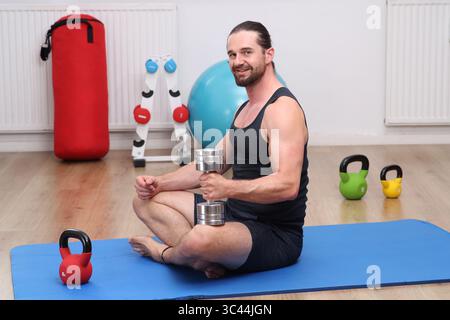 Weißer Mann mit Bart, der mit Hantel auf dem Boden im Fitnessstudio sitzt, schwarzes Hemd Stockfoto