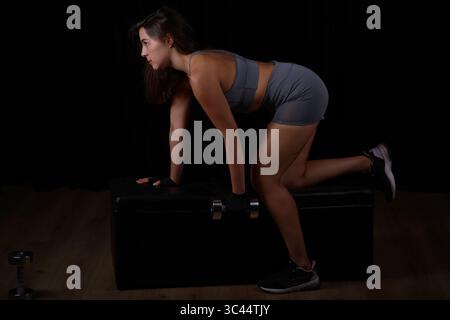 Junge Frau, die mit Hantel im Fitness-Outfit auf der Bank kniend trainiert Stockfoto