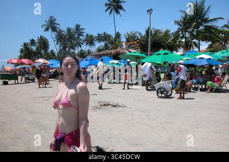 Eine Frau in einem rosafarbenen Bikini steht am Strand, mit anderen Leuten und Sonnenschirmen im Hintergrund. Stockfoto