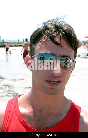 Ein Mann mit Sonnenbrille und rotem Tanktop ist am Strand. Die Reflexion in seiner Sonnenbrille zeigt andere Menschen am Strand. Stockfoto