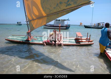 Ein Paar sitzt auf einem kleinen Boot mit einem Segel im Wasser. Andere Boote befinden sich im Hintergrund, und ein Mann steht im Wasser in der Nähe eines anderen Bootes. Stockfoto