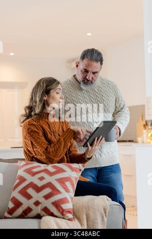 Seniorenpaar interagiert mit dem Tablet, während er zu Hause auf dem Sofa sitzt und Kaffeetasse in der Nähe der Küche hält Stockfoto
