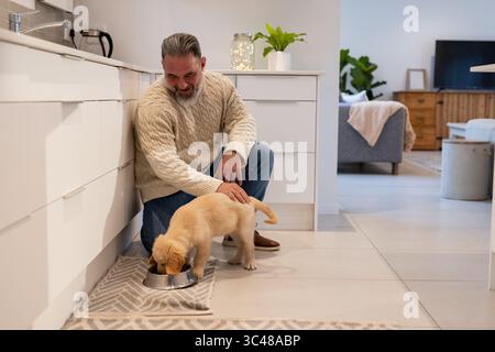 Mann kniend klopfender Welpe, der aus einer Edelstahlschale auf geometrischer Matte in der Küche zu Hause isst Stockfoto