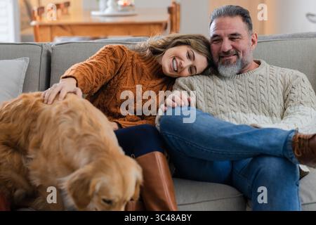 Ein Paar sitzt zu Hause auf dem Sofa und trägt Strickpullover und Stiefel, die Golden Retriever streicheln Stockfoto