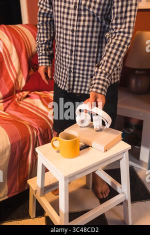 Ein Mann hält kabellose Kopfhörer in der Nähe eines Buches und einer Kaffeetasse auf einem kleinen Tisch, um sich auf einen entspannenden Moment zu Hause vorzubereiten Stockfoto