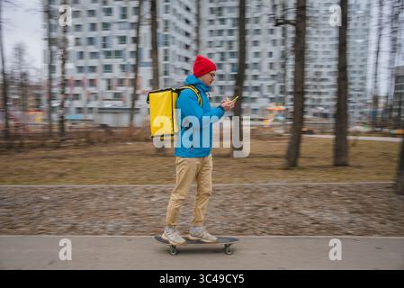 Ein Mann, der mit einer Smartphone-App auf einem Skateboard unterwegs ist, durch einen Stadtpark, der von Wohnhäusern umgeben ist, navigiert und moderne Bequemlichkeit und Effizienz bietet Stockfoto