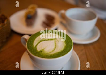 Warmer Matcha Latte auf einem Holztisch mit zartem Herzdesign in Milchschaum, umgeben von einem verschwommenen Hintergrund aus Kuchen und einer weiteren Tasse, sorgt für eine gemütliche Café-Atmosphäre Stockfoto