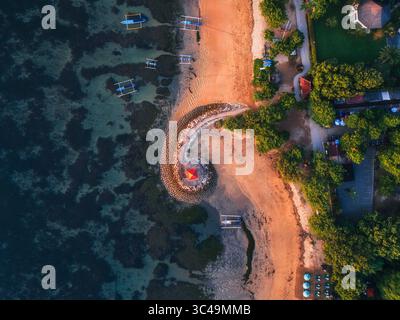 Aus der Vogelperspektive auf einen spiralförmigen Steg, der sich ins Meer erstreckt, wo der dunkle Ozean auf das goldene Ufer trifft, gesäumt von lebhaften grünen Bäumen, Sanur, Bali, Indonesien. Stockfoto