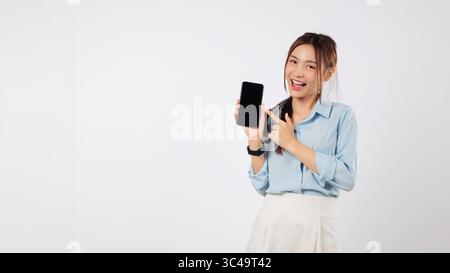 Eine glückliche asiatische Frau zeigt ihr Handy und zeigt mit dem Finger auf das Display Stockfoto