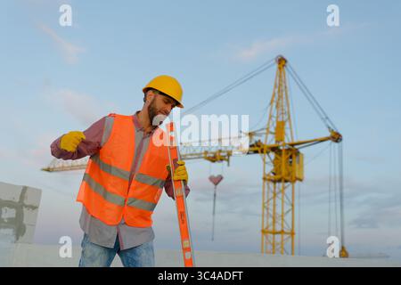 Bauarbeiter, der eine Warnweste anzieht und die Wasserwaage auf der Baustelle hält, mit Kran im Hintergrund Stockfoto