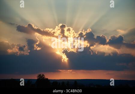 Die Sonne geht hinter dramatischen Wolken unter und wirft goldene Lichtstrahlen aus. Der Himmel ist in warmen Orange- und Rosa-Tönen gemalt, im Kontrast zu dunkleren Wolken. Silhouettenbäume und weit entfernte Häuser verleihen der Abendszene Tiefe. Stockfoto
