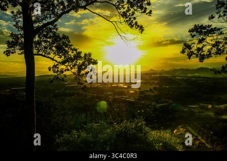 Malerischer Blick in die Dämmerung vom Berggipfel aus mit Blick auf das Dorf, Reisfelder und tropischen Wald unter einem leuchtenden goldenen Himmel. Silhouettenbäume im Vordergrund. Stockfoto