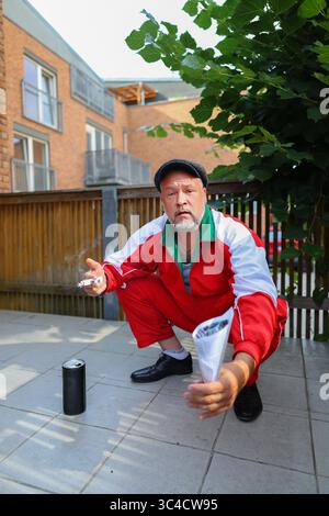 Ernster Mann in rotem Trainingsanzug, der draußen hockt, Zigarette hält, urbaner Parody Charakter. Stockfoto