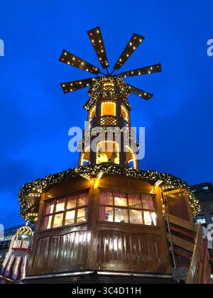 Eine kunstvoll verzierte, mehrstöckige hölzerne Weihnachtspyramide, die mit zahlreichen warmweißen Lichtern und grünen Girlanden vor einem tiefblauen Himmel geschmückt ist Stockfoto
