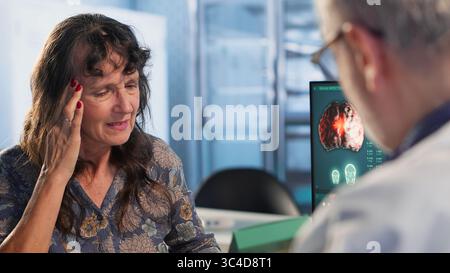 Alte Frau, die mit einer schweren Migräne zu kämpfen hat und um Medikamente bittet, Arzt, der einen Erholungsplan mit starken Schmerzmitteln aufstellt, um Schmerzen beim Kontrollbesuch zu lindern. Kamera B.. Stockfoto
