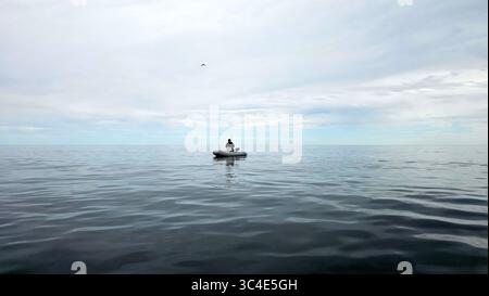 Eine Einzelfigur in einem kleinen aufblasbaren Boot schwimmt auf der weiten, ruhigen Weite des Meeres unter einem bewölkten Himmel, mit einem Seevögel (möglicherweise eine Möwe oder Ter) Stockfoto