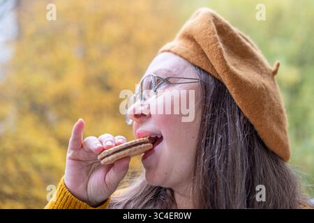 Reife hungrige Frau beißt Sandwichkekse mit lustigem Verlangen Ausdruck, Comic Gesicht Ausdruck sehnsüßen Snack, draußen in der Herbstsaison, gesund Stockfoto