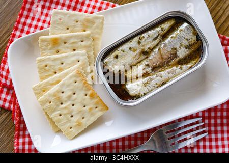 Nahaufnahme einer offenen Dose Sardinen in Öl, serviert neben einem Haufen einfacher quadratischer Cracker auf einer rechteckigen weißen Platte. Stockfoto