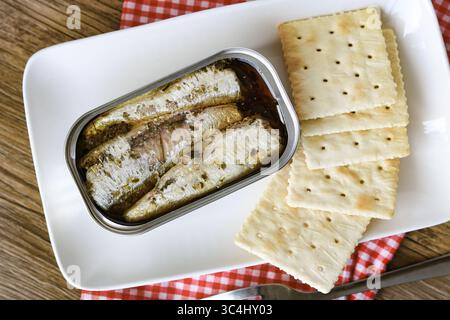 Nahaufnahme einer offenen Dose Sardinen in Öl, serviert neben einem Stapel einfacher quadratischer Cracker auf einer rechteckigen weißen Platte. Stockfoto