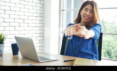 Frau streckt den Arm hoch und sitzt falsch in der Position des Home Office-Schreibtisches. Rückseite einer jungen asiatischen Frau müde von der Arbeit Körper Stress Rücken Schmerz Büro Sy Stockfoto