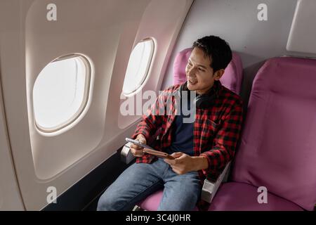 Ein junger Mann sitzt in einem Flugzeug und genießt seinen Flug, während er sein Smartphone und seinen Reisepass überprüft. Er wirkt entspannt und zufrieden und spiegelt die Moderne wider Stockfoto