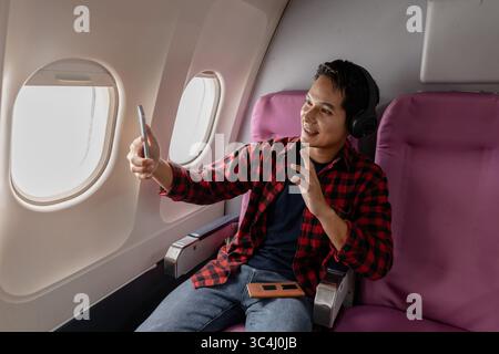 Ein fröhlicher junger Mann genießt seinen Flug, während er sein Smartphone benutzt und Kopfhörer trägt. Er sitzt am Flugzeugfenster und zeigt Aufregung und Happy Stockfoto