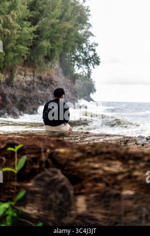 Rückansicht eines jungen Mannes mit Rucksack, der auf Treibholz sitzt und Wellen unter den Klippen in der Nähe von Hilo, Big Island, Hawaii, an einem bewölkten Tag beobachtet. Stockfoto
