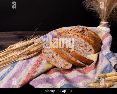 Ein rustikaler Brotlaib auf einem farbenfrohen Leinentuch, begleitet von Weizenohren, schafft eine warme und einladende Atmosphäre Stockfoto