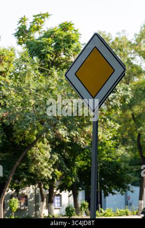 Ein gelbes, rautenförmiges Straßenschild mit weißem Rand, umgeben von üppig grünen Bäumen und einem Gebäude in städtischer Umgebung Stockfoto
