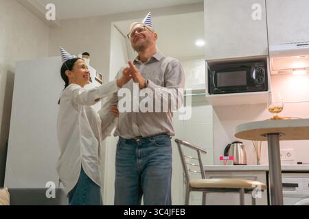 Geburtstagsfeier für Paare Home Party: Fröhliches reifes Paar tanzt fröhlich in ihrer Küche und feiert mit Partyhüten einen Geburtstag. Stockfoto