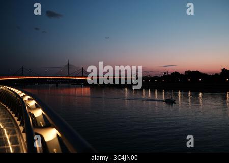 Wunderschöner Sonnenuntergang über der Save in Belgrad mit beleuchteter Brücke und einem kleinen Boot über das Wasser. Friedliche urbane Landschaft. Stockfoto