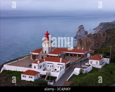 Blick aus der Vogelperspektive auf einen markanten Leuchtturm mit einer leuchtenden roten Decke über zerklüfteten Klippen und dem riesigen Atlantik, Colares, Lisboa, Portugal. Stockfoto