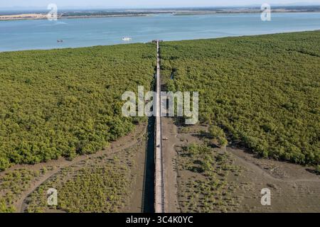 Aus der Vogelperspektive einer langen, geraden Straße führt durch einen üppigen grünen Wald in Richtung des schimmernden blauen Meeres, Teknaf, Chittagong Division, Bangladesch. Stockfoto