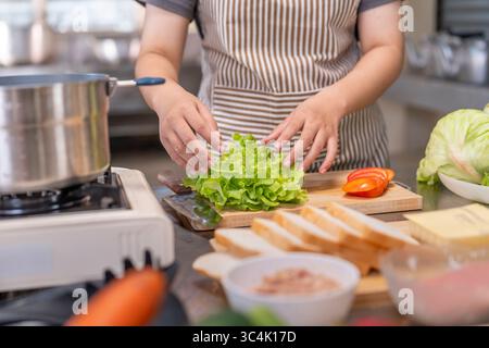 Eine Küchenchefin in einer Schürze arrangiert sorgfältig frischen grünen Salat auf einem hölzernen Schneidebrett mit anderen Zutaten. Ideal für gesunde Ernährung, Lebensmittelvorbereitung Stockfoto