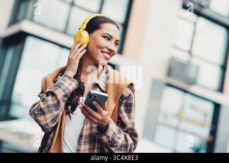Junge Frau mit gelben Kopfhörern, die Musik mit dem Telefon in der Hand genießt, während sie durch die sonnige Stadtlandschaft spaziert Stockfoto