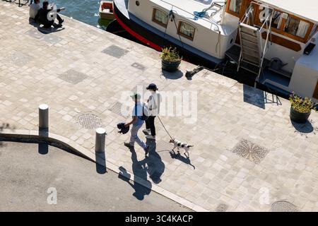 Hochwinkelansicht eines Paares, das mit seinem Hund an einem sonnendurchfluteten Pflasterkai spaziert. In der Nähe liegt ein Kanalboot. Ideal für Reisen, lifestyl Stockfoto