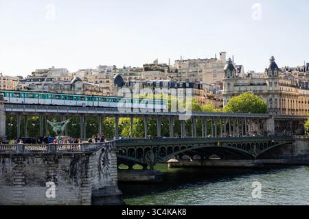 Eine Pariser Brückenszene: Eine türkisfarbene U-Bahn durchquert ein historisches Gebäude, Fußgänger schlendern entlang des Gehwegs und elegante Pariser Gebäude bilden sich Stockfoto
