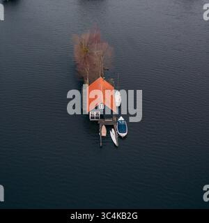 Blick aus der Vogelperspektive auf ein einsames Haus mit einem leuchtenden orangefarbenen Dach inmitten des dunklen Wassers, ein Leuchtfeuer der Wärme in der gedämpften Landschaft, Breukelen, Utrecht, Niederlande. Stockfoto