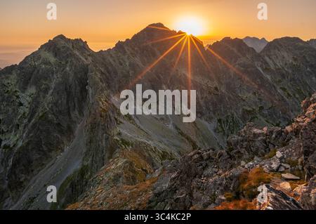 Aus der Vogelperspektive auf die strahlende Sonne über felsigen Gipfeln, die lange Schatten wirft und das zerklüftete Gelände, Vysoké Tatry, Region Prešov, Slowakei, hervorhebt. Stockfoto